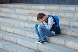 Abbildung zeigt einen jungen Mann/Jugendlichen auf einer Treppe sitzend, das Gesicht in die Hände gestützt Jugendliche 2.png